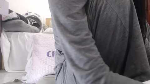 Media: Video of a person in a gray sweatshirt, sitting on a bed with a white quilted pillow and a partially visible black and white washing machine in the background.