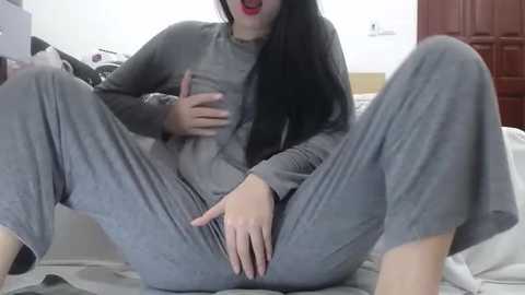 Media: Video of an Asian woman with long black hair, wearing a gray sweatshirt and matching sweatpants, sitting with legs spread, exposing her crotch, in a bedroom.