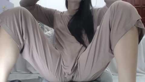 Media: Video of an Asian woman with long black hair, wearing a beige, long-sleeved onesie, sitting with legs spread, indoors on a bed.