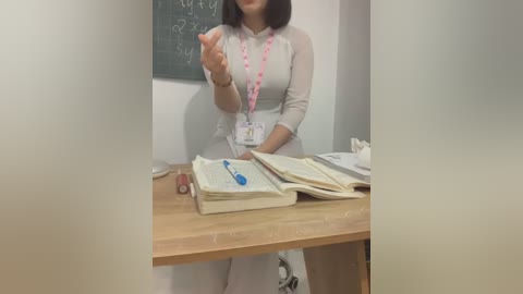 Media: Video of a woman in a beige dress and pink lanyard, standing at a wooden table with open books and papers, in a classroom with a chalkboard in the background.