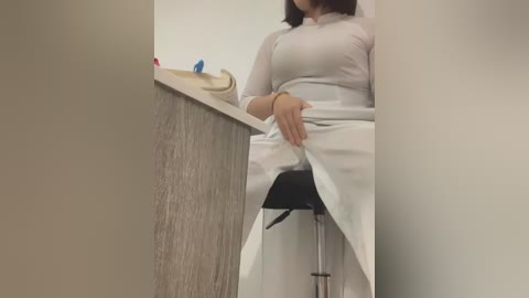 Media: A video of a woman in a white lab coat, seated at a desk with a beige clipboard, a blue pen, and a black stool. She is partially obscured by a beige wall.