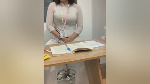 Media: Video of a woman with medium-length dark hair, wearing a light beige long-sleeve shirt, seated at a wooden desk, with an open notebook, pen, and a yellow highlighter on the desk.