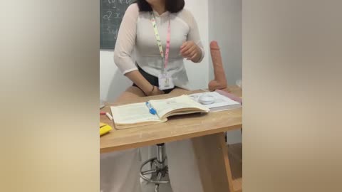 Media: A video shows an Asian woman with shoulder-length black hair, wearing a sheer white blouse and black shorts, sitting at a wooden desk, holding an erect penis and reading a book.