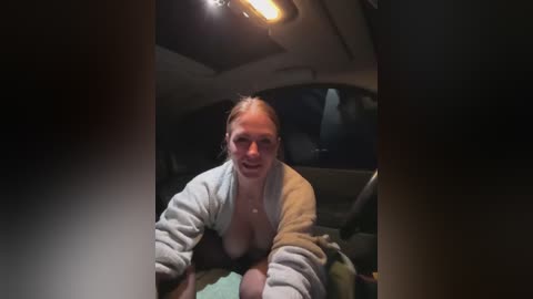 Media: Video of a blonde woman in a beige robe, sitting in a dimly lit car, smiling, with a partially open window and a blurred background.