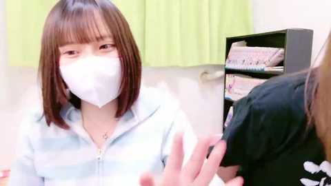 Media: Video of a young woman with straight, shoulder-length brown hair and light skin wearing a white mask and light blue hoodie, holding a black shirt with a white logo, in a brightly lit room with green curtains and a black shelf filled with folded clothes.