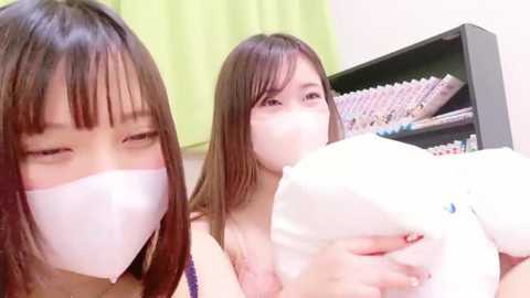 Media: Video of two young Asian women, both wearing white surgical masks, hugging a large white teddy bear in a brightly lit room with green curtains and a bookshelf in the background.