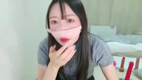 Media: A video of an Asian woman with long black hair and pale skin, wearing a pink face mask, a gray shirt, and sitting in a bedroom with a white bed and pink sex toys.