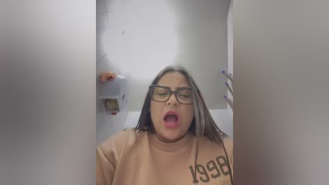 Media: Video of a woman with long, straight brown hair, wearing glasses, a beige sweatshirt with \"1998\" on the chest, and a surprised expression in a dimly lit, narrow hallway.