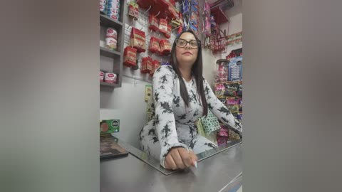 Media: Video of a female shopkeeper with long black hair, wearing glasses and a white floral-patterned dress, standing behind a glass display counter in a brightly lit convenience store.