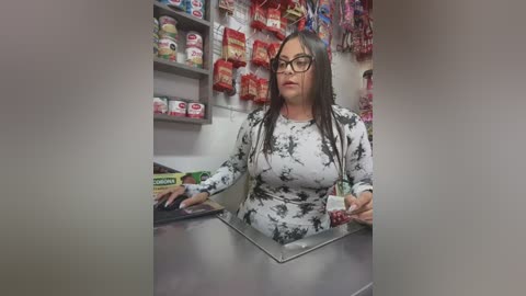 Media: Video of a middle-aged woman with long dark hair and glasses, wearing a white and black tie-dye top, standing behind a candy store counter. Background features colorful candy displays and shelves.