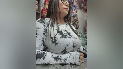 Media: Video of a plus-sized woman with long dark hair, wearing a white tie-dye long-sleeve top and glasses, standing in a grocery store aisle filled with colorful snack bags.