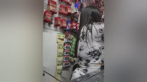 Media: Video of a woman with long black hair, wearing a white and black floral dress, standing in a brightly lit grocery aisle. Red and green snack bags line the shelves behind her.