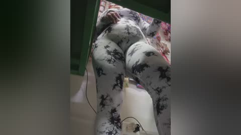 Media: Video of a plus-sized woman with large breasts and wide hips, wearing tight, black-and-white tie-dye leggings, sitting on a white toilet in a dimly lit bathroom.