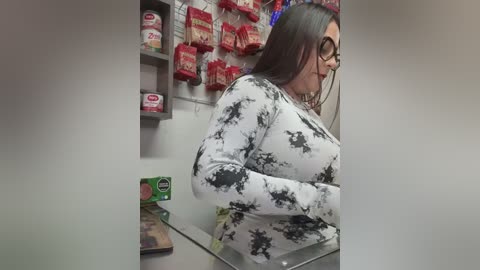 Media: A video of a plus-sized woman with long brown hair, wearing black-rimmed glasses, a tight white floral-patterned top, and standing behind a glass display case in a convenience store.