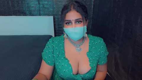 Media: Video of a South Asian woman with medium skin tone, wearing a green dress with a deep V-neck, revealing ample cleavage. She has a light blue face mask, dark hair, and a gold necklace. Background shows a dark wall and a bed with a gray sheet.