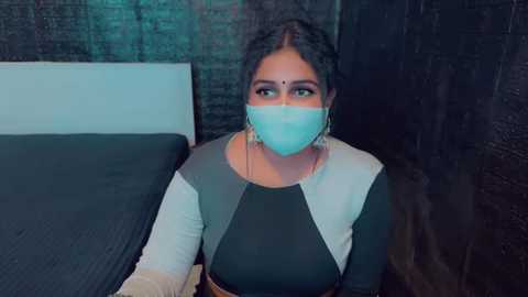 Media: Video of a South Asian woman in a black and white saree, with a teal mask and bindi, sitting on a bed in a dimly lit room with dark brick walls.