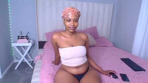 Media: Video of a slender, dark-skinned woman with an orange headwrap, wearing a strapless pink crop top and black panties, sitting on a pink bed with white headboard, holding a phone.