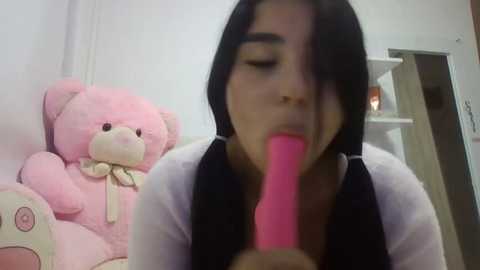 Media: Video of a young woman with long black hair, wearing a white shirt, licking a pink dildo. Background includes a plush teddy bear and a white doorframe.