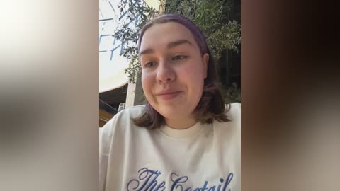 Media: Video of a smiling young woman with light skin, brown hair, and a white T-shirt with blue text, standing outdoors near a tree and yellow structure.