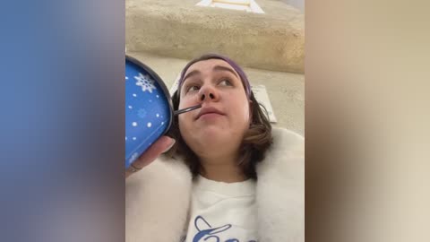 Media: A video of a young woman with short brown hair and a purple headband, wearing a white shirt, having a blue snowflake-topped cone applied to her nose.