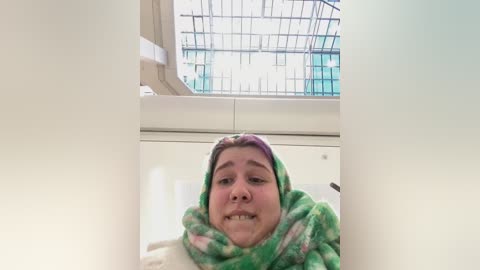 Media: A video shows a middle-aged woman with light skin and a round face, wearing a green and pink patterned headscarf and a beige sweater, standing indoors near a large glass window with a metal grid.