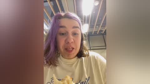 Media: Video of a young woman with shoulder-length, purple-dyed hair, wearing a white t-shirt, sitting in a dimly lit, modern interior with exposed wooden beams and industrial lighting.