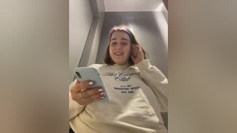 Media: Video of a young woman with light skin and long brown hair, wearing a beige sweatshirt with text, taking a selfie in a narrow, beige-walled hallway.