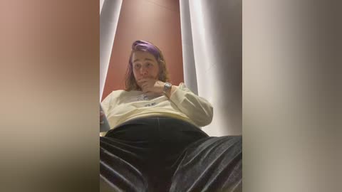 Media: A video of a person in a low-angle view, wearing a yellow sweatshirt and black pants, seated in a white-walled stall, with a purple headband.