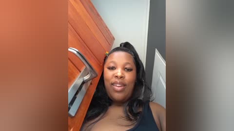 Video of a smiling Black woman with long black hair in a high ponytail, standing in a bathroom with a wooden door and silver handle. She wears a black top, and the background includes a white door and gray walls.