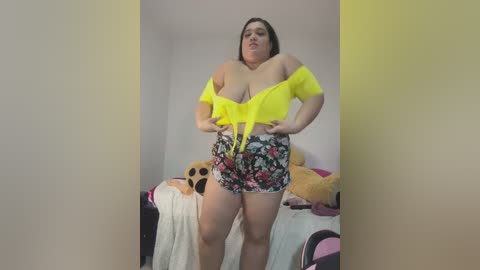 A video of a plus-size woman with fair skin and brown hair, wearing a neon yellow off-shoulder crop top and floral shorts, standing in a bedroom with stuffed animals and a bed.