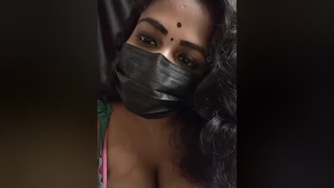 Media: Video of a South Asian woman with medium brown skin, long black curly hair, and a black mask covering her nose and mouth, wearing a green and pink top, with a dark background.