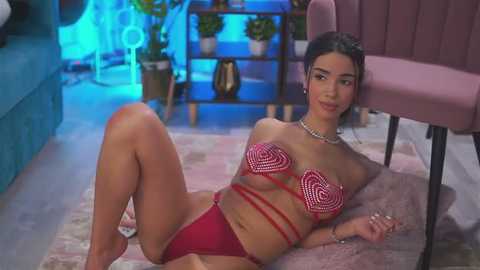 Video of a slim, tan-skinned woman with dark hair, wearing a red and white heart-patterned bikini, lying on a plush rug in a modern room with pink chairs and blue lighting.