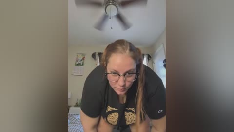 Media: Video of a Caucasian woman with glasses, light brown hair in pigtails, wearing a black t-shirt with a graphic design, leaning forward in a bedroom with a ceiling fan and framed artwork on beige walls.