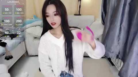 Media: Video of a young Asian woman with long black hair, red lipstick, and white sweater, sitting on a white couch in a dimly lit living room. She is holding a pink object.