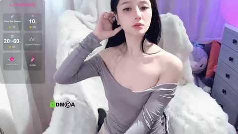 Media: Video of an Asian woman with pale skin, long black hair, and small breasts, wearing a grey off-shoulder sweater, sitting on a white fur-covered bed. Background includes a dresser, fluffy white pillows, and a dimly lit room.