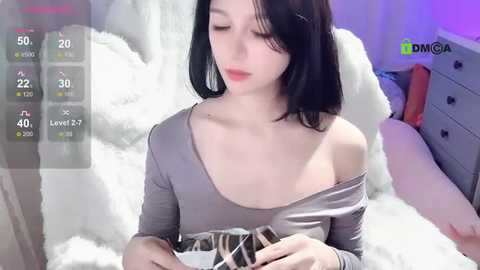 Media: A video of a young Asian woman with fair skin and straight black hair, wearing a gray off-shoulder top, lying on a white fur blanket in a cozy bedroom.