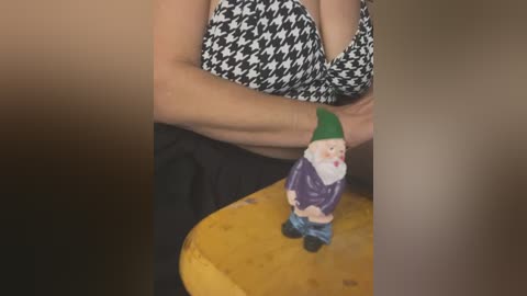 Video of a person with fair skin wearing a black-and-white houndstooth top and blue jeans, holding a colorful ceramic elf figurine with a green hat and red nose.