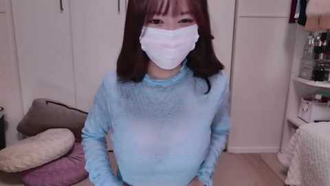 Media: Video of an Asian woman with long black hair and fair skin wearing a blue long-sleeve shirt, white mask, and purple face shield, standing in a cluttered bedroom with white cabinets, a bed, and various items.