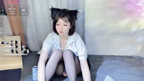 Media: A young Asian woman with short black hair, wearing a white shirt and grey tights, sits on a gray rug, looking downcast. The background features a white curtain and a cardboard box.