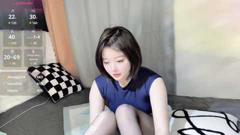 Media: Video of a young Asian woman with short black hair, wearing a blue sleeveless top and white tights, sitting on a gray couch in a modern room with gray and white curtains.