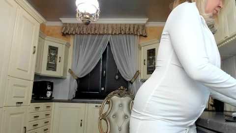 Media: Video of a curvy woman in a white long-sleeve top, leaning over a kitchen counter, with a fancy chair and ornate curtains in the background.