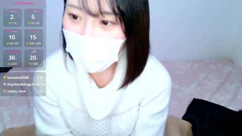 Media: Video of a young East Asian woman with straight black hair, wearing a white mask and a white sweater, sitting on a bed.