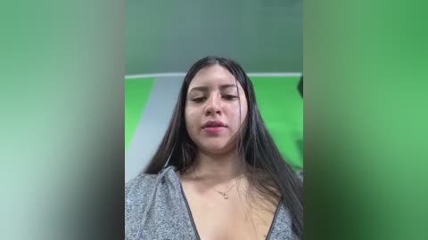 Media: Video of a young Latina woman with long, straight black hair, wearing a gray V-neck sweater, standing in a bright, green-lit room. She has a neutral expression and is slightly out of focus.