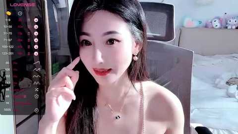 Media: A video of an East Asian woman with long black hair, fair skin, and red lipstick, wearing a delicate necklace. She sits on a bed with a plush toy and a white blanket in the background.