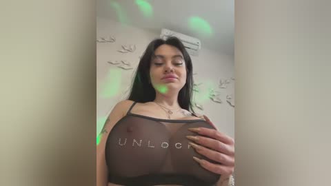 Media: A video of a woman with long black hair, wearing a sheer black top with \"UNLOOOK\" printed on it, revealing her large breasts, in a bedroom with minimalist decor.