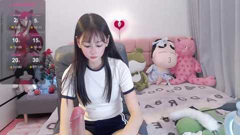 Media: Video of an East Asian woman with long black hair in a white T-shirt, seated at a desk. She holds a large pink dildo. Background features a plush teddy bear and a pink heart light.