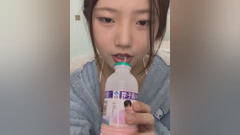 Media: A video of an Asian woman with light skin, straight black hair, wearing a blue bathrobe, drinking from a white plastic bottle with a purple label, indoors with a blurred background.