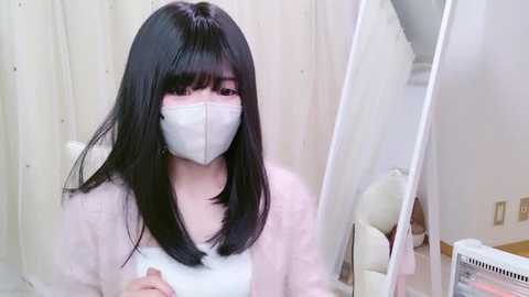 Media: Video of an East Asian woman with long black hair and pale skin wearing a white mask and light sweater, sitting in a room with white curtains and a white fan.