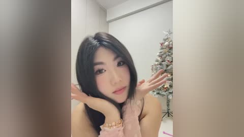 Media: A video of an Asian woman with straight black hair, fair skin, and makeup, posing in front of a decorated Christmas tree in a softly lit room. She wears a pink sweater and has a playful expression.