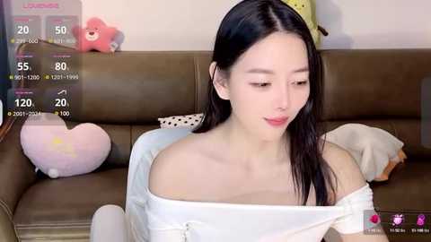 Media: Video of a slender East Asian woman with long black hair, wearing a white off-shoulder top, sitting on a brown leather couch. Background features a teddy bear and heart-shaped pillow. Live stream overlay on the left.